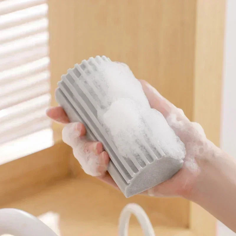 Hand holding a gray scrubber with soap suds against a neutral background
